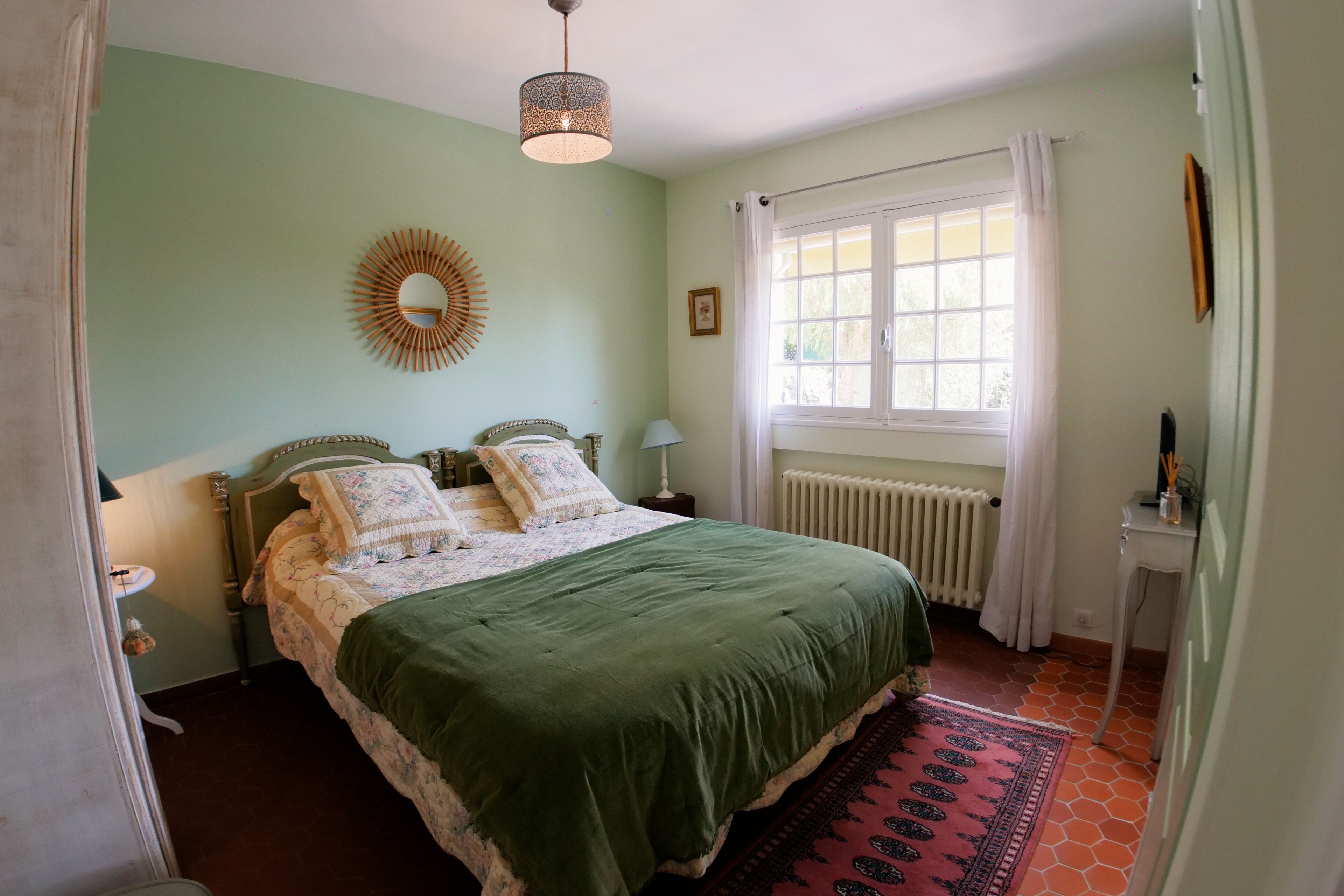 A bedroom with a bed, a bedside table, a lamp, a window with curtains, a radiator, and a wall-mounted mirror.