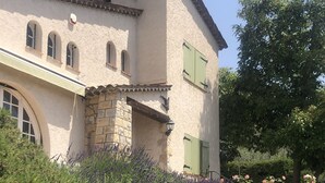 A two-story stone building with a sloped roof, green shutters, and a stone staircase leading to the entrance.