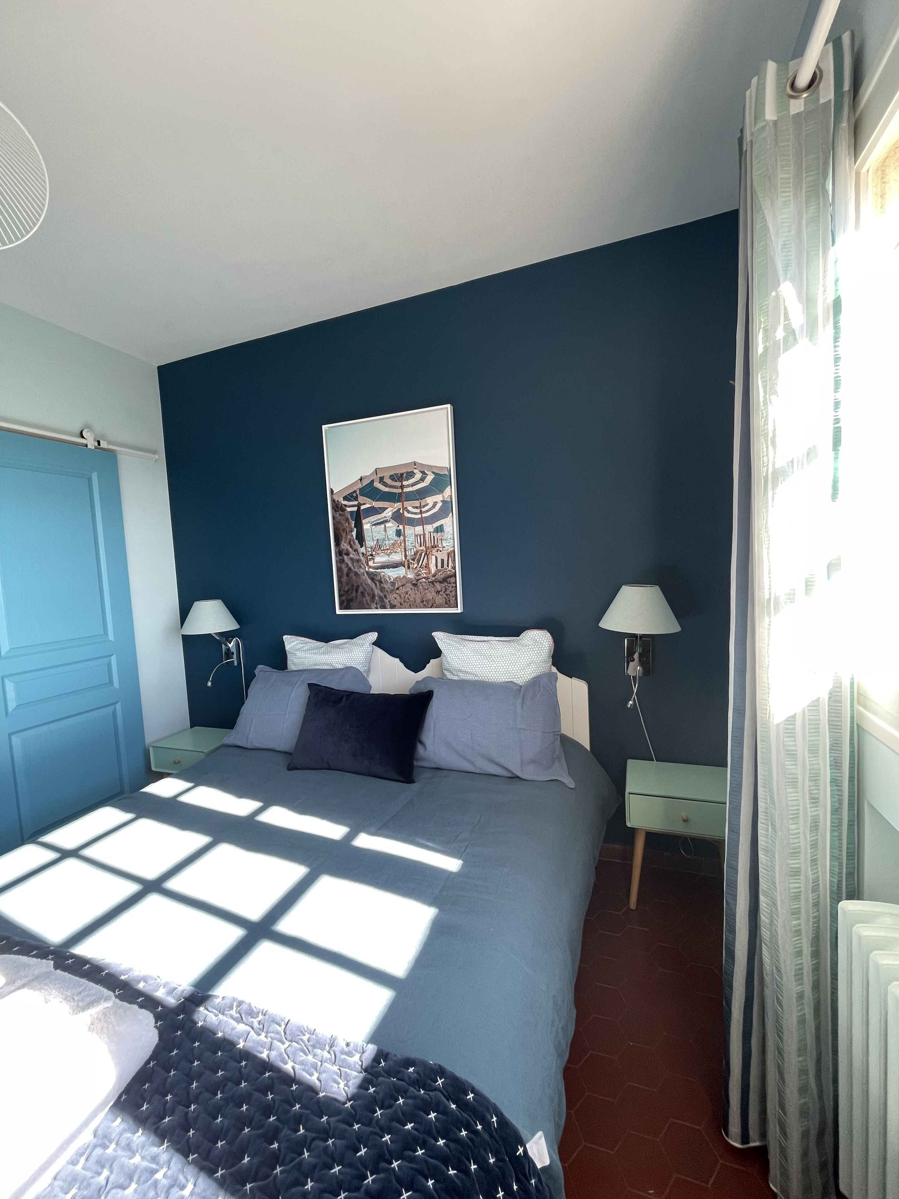 Un dormitorio con una cama, dos mesitas de noche, una lámpara de pared, un cuadro y una ventana con cortinas.