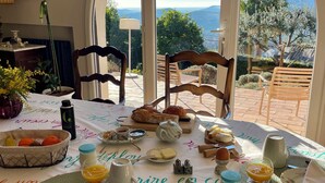 Une table de petit-déjeuner dressée avec divers aliments, et une vue sur l’extérieur grâce à de grandes fenêtres.