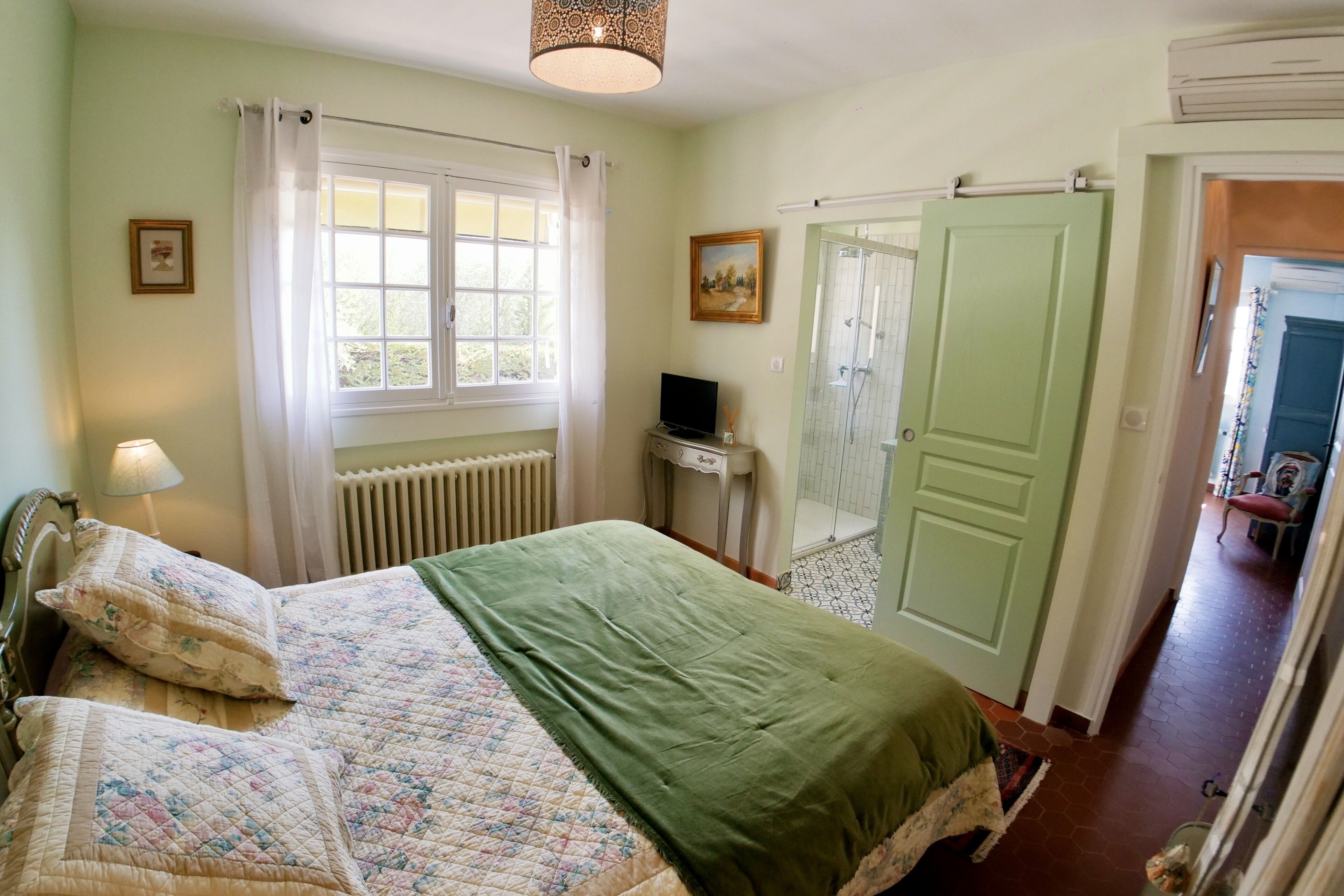 A bedroom with a bed, a window with curtains, a radiator, a television, and a door leading to another room.
