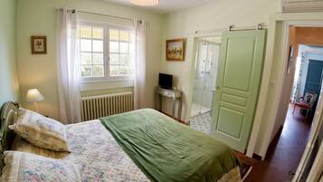 A bedroom with a bed, a window with curtains, a radiator, a television, and a door leading to another room.
