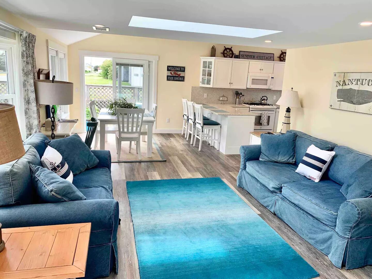 A modern living room with a blue sofa, a wooden coffee table, and a kitchen area with white cabinets and a dining table.