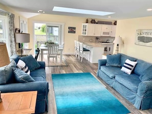 Una sala de estar moderna con un sofá azul, una mesa de centro de madera, y una zona de cocina con armarios blancos y una mesa de comedor.