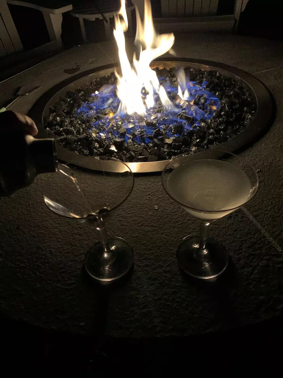 A fire pit with flames, two wine glasses, and a spoon on a textured surface.