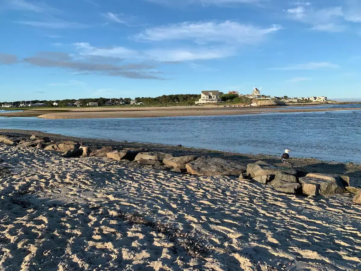 A sandy beach with a rocky breakwater and a view of houses across the water.
