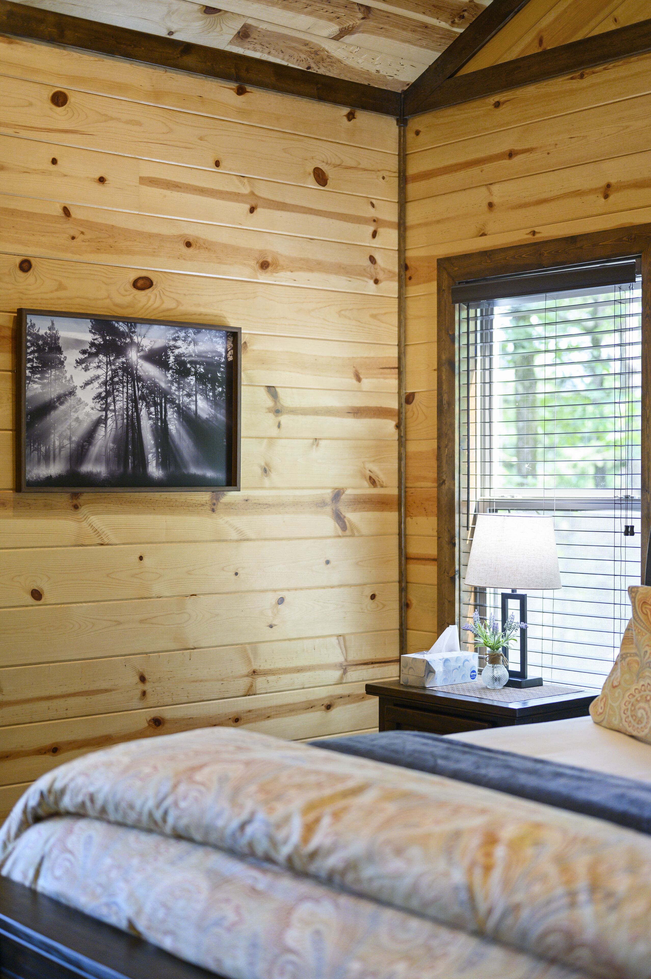 A wooden cabin bedroom with a bed, a framed forest photograph, a nightstand with a lamp, and a window with blinds.
