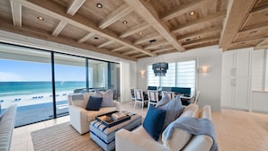 A spacious living room with a large window overlooking a beach, wooden ceiling beams, and a dining area with white furniture.