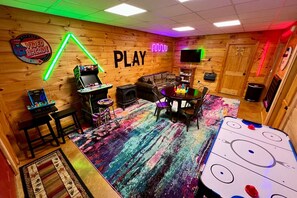A room with wooden walls, a neon 'PLAY' sign, a video arcade, a couch, a table with chairs, and an ice hockey table.