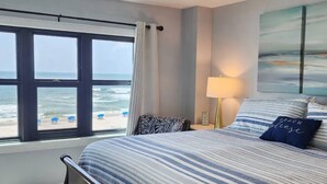 Ein Schlafzimmer mit großem Fenster mit Blick auf den Strand, einem Bett mit gestreifter Bettwäsche, einer Nachttischlampe und einem an der Wand hängenden Gemälde.