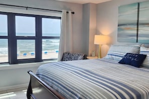 A bedroom with a large window overlooking the beach, a bed with striped bedding, a bedside lamp, and a wall-mounted painting.