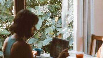 Una persona sentada a una mesa con una computadora portátil y una taza, con vista a follaje verde a través de una ventana.