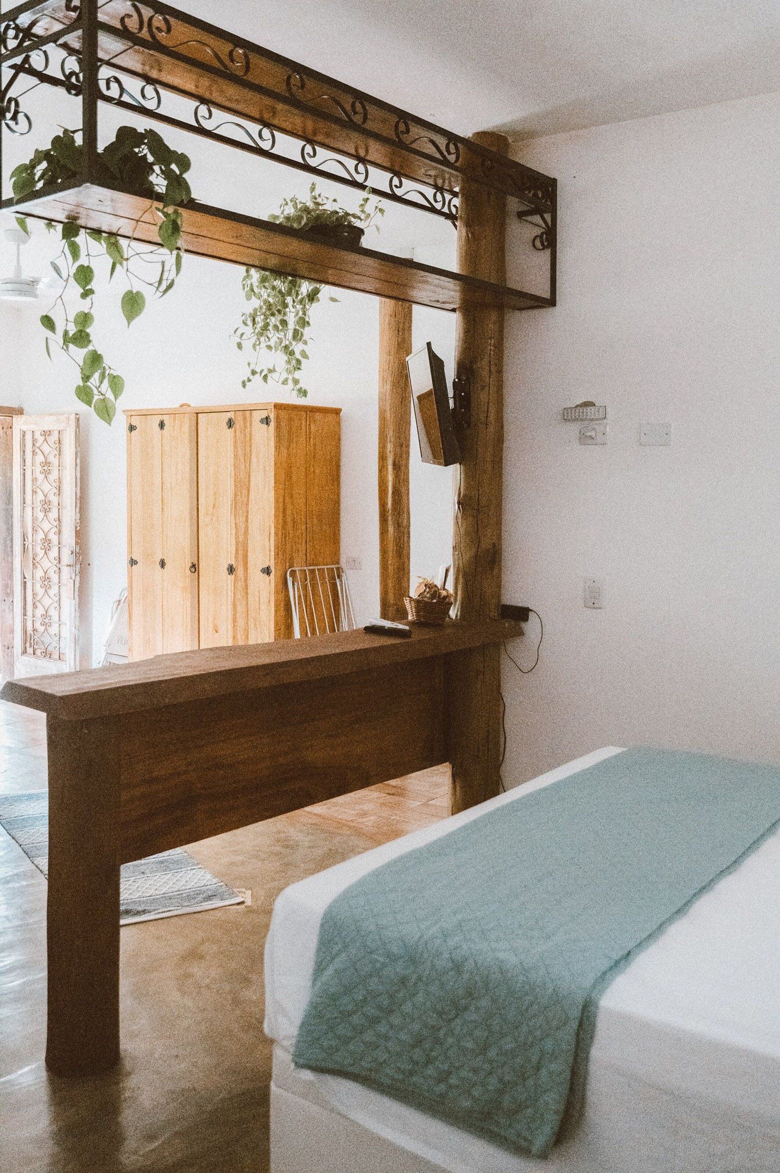 Une chambre à coucher avec un lit, un bureau en bois, une plante suspendue, et une vue sur une armoire en bois et une chaise.