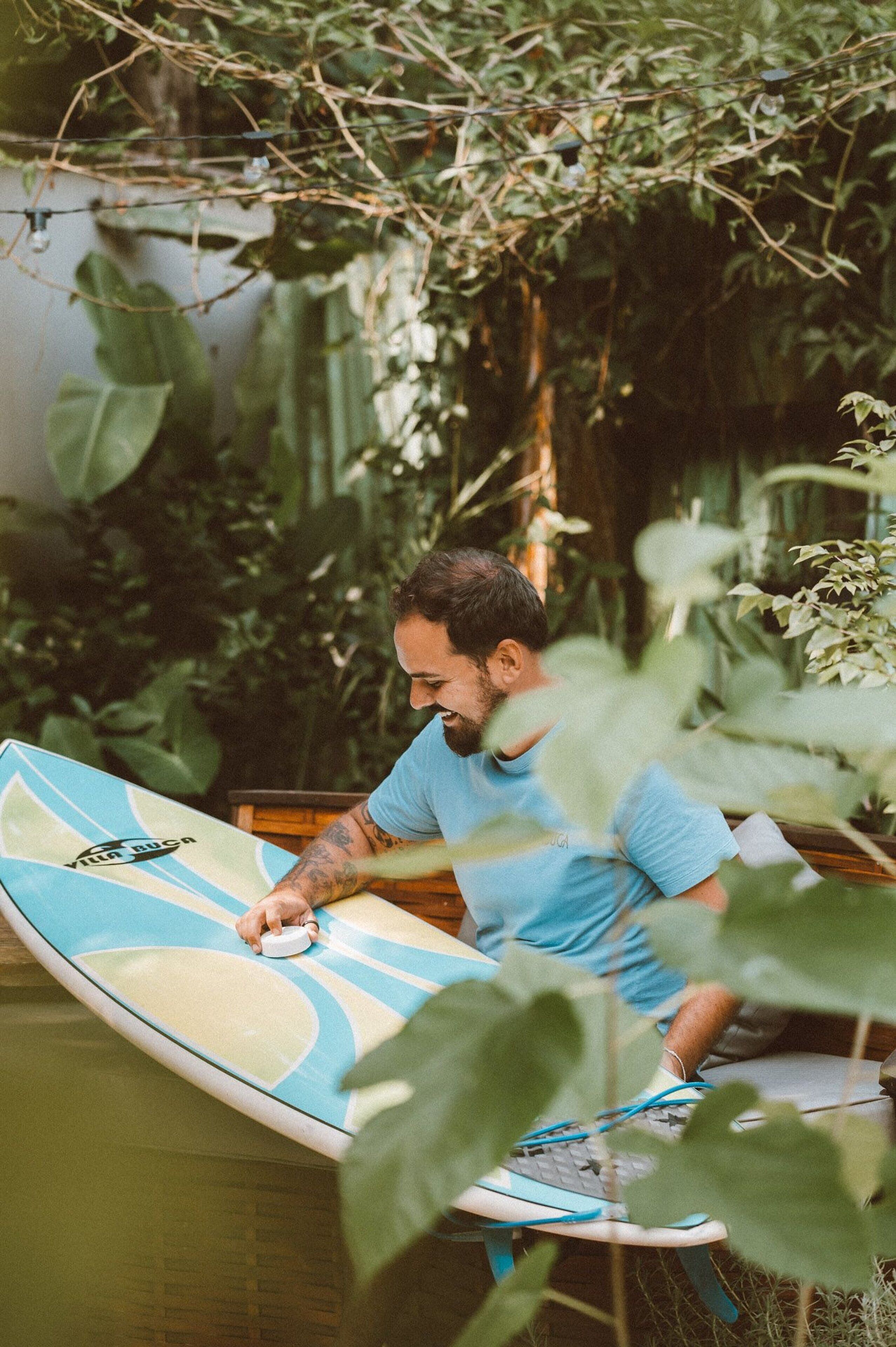 Una persona sentada en una silla con una tabla de surf cerca, rodeada de follaje.