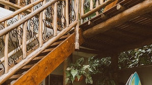 Eine Holztreppe mit verzierten Metallgeländern, eine Hängematte und Surfbretter in einem überdachten Außenbereich mit Topfpflanzen.