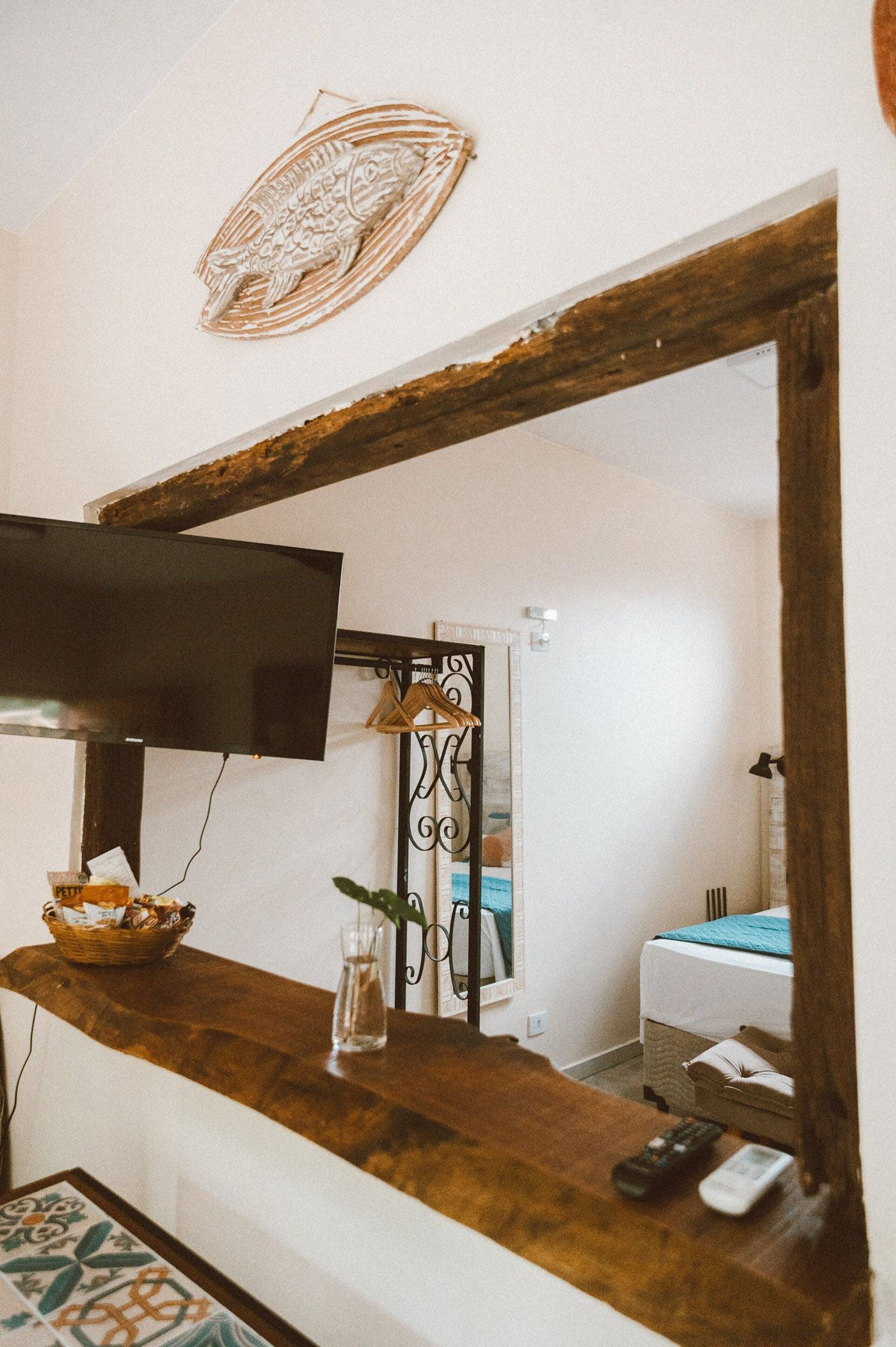 A hotel reception area with a wooden counter, a television, a decorative wall piece, and a mirror reflecting a room with a bed and a door.