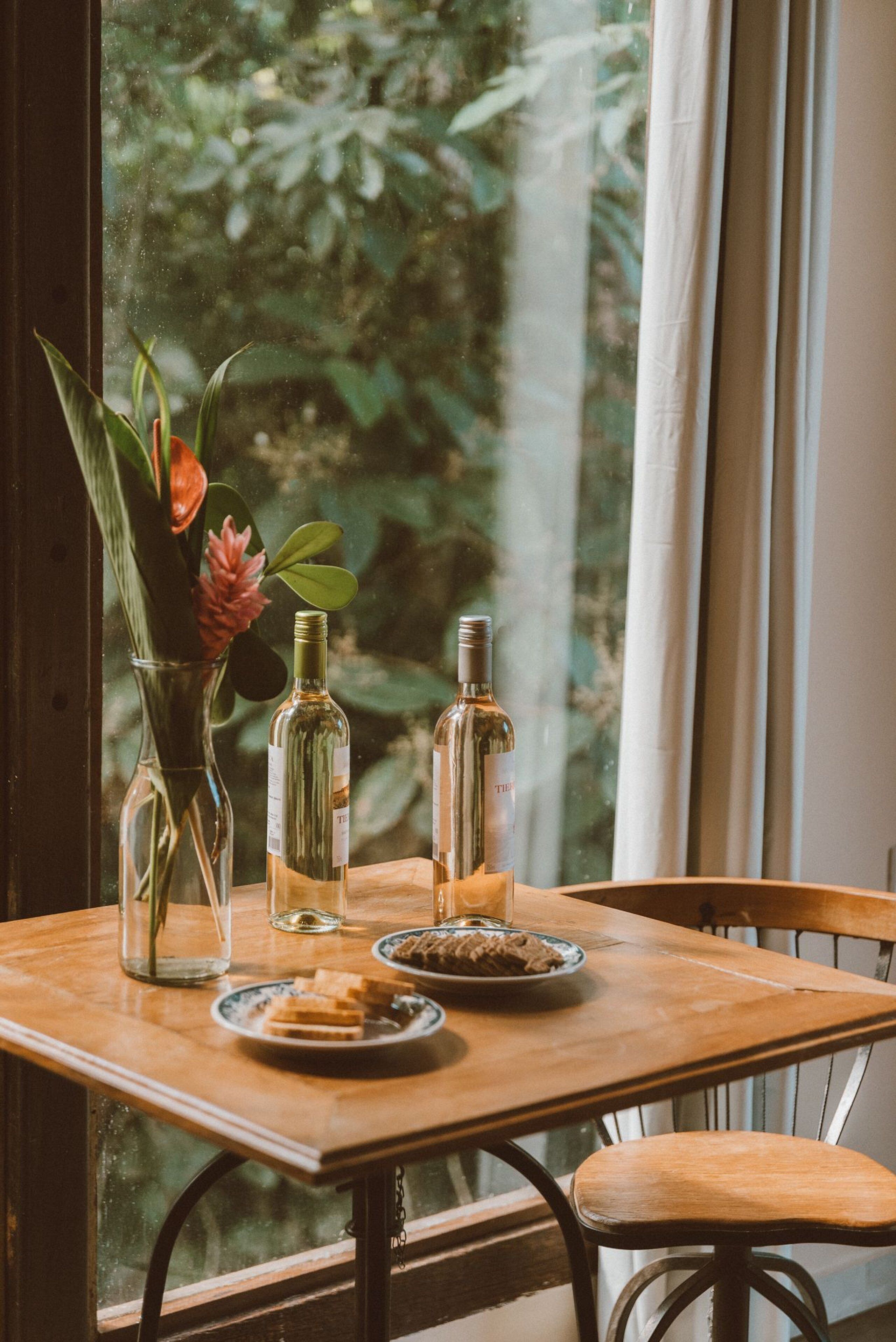 Una mesa de madera con un jarrón de flores, dos botellas y un plato de comida junto a una ventana con vistas a follaje.