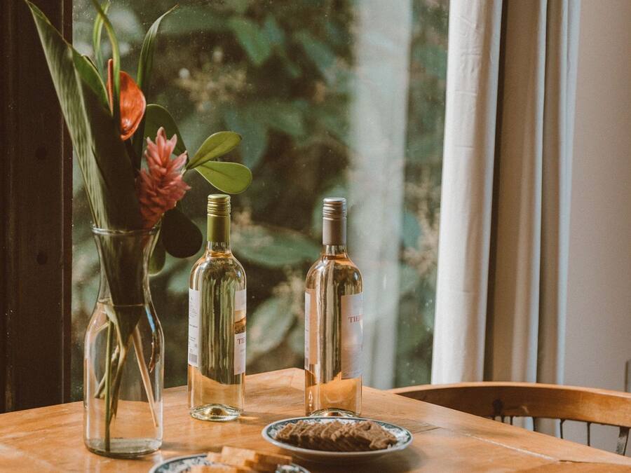 Una mesa de madera con un jarrón de flores, dos botellas y un plato de comida junto a una ventana con vistas a follaje.