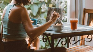 Eine Person sitzt an einem Tisch mit einer Schüssel Müsli und einem Glas Saft, im Hintergrund ein Fenster.