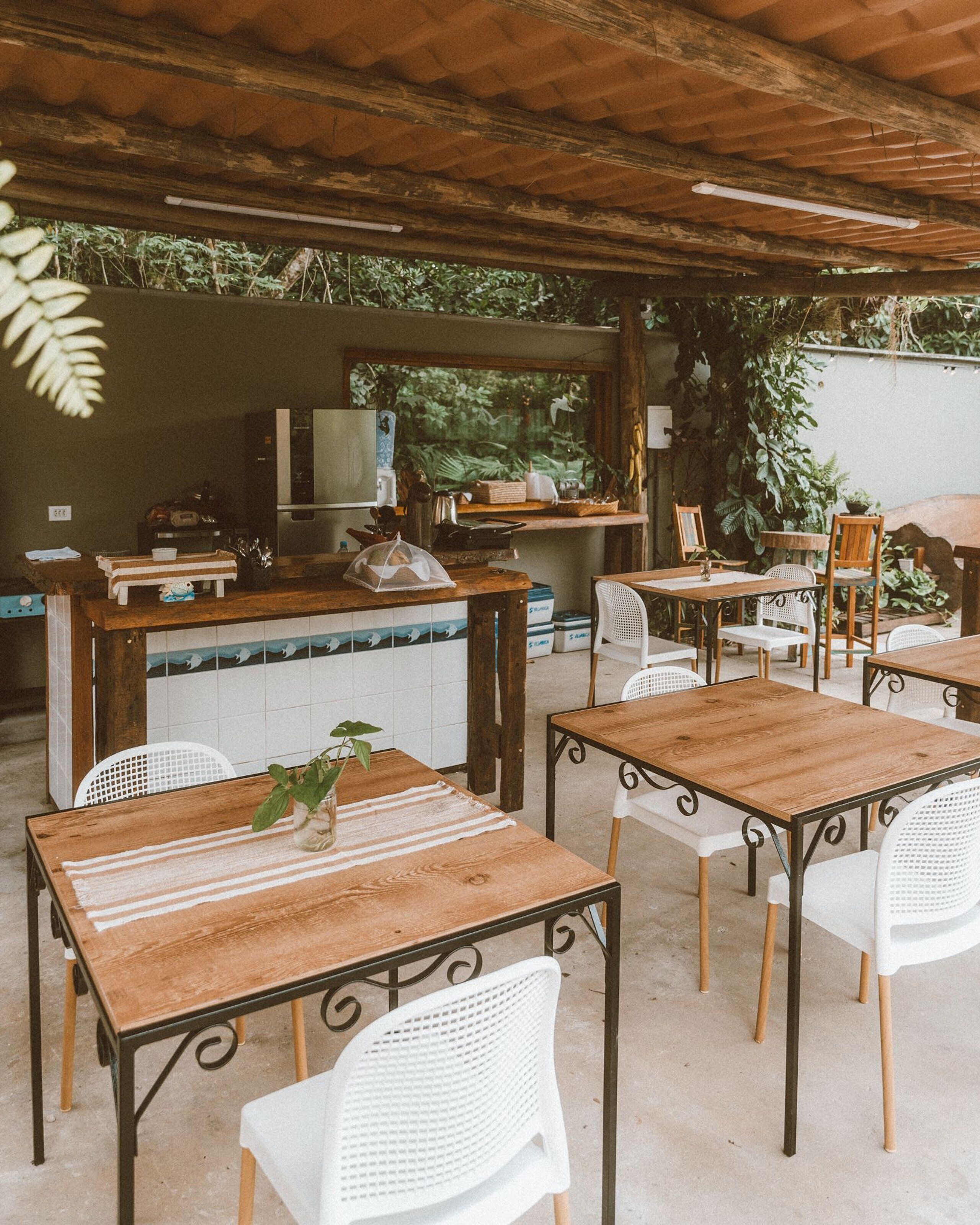 Un patio exterior con mesas de madera y sillas blancas, un salpicadero de azulejos y diversas plantas y electrodomésticos de cocina.