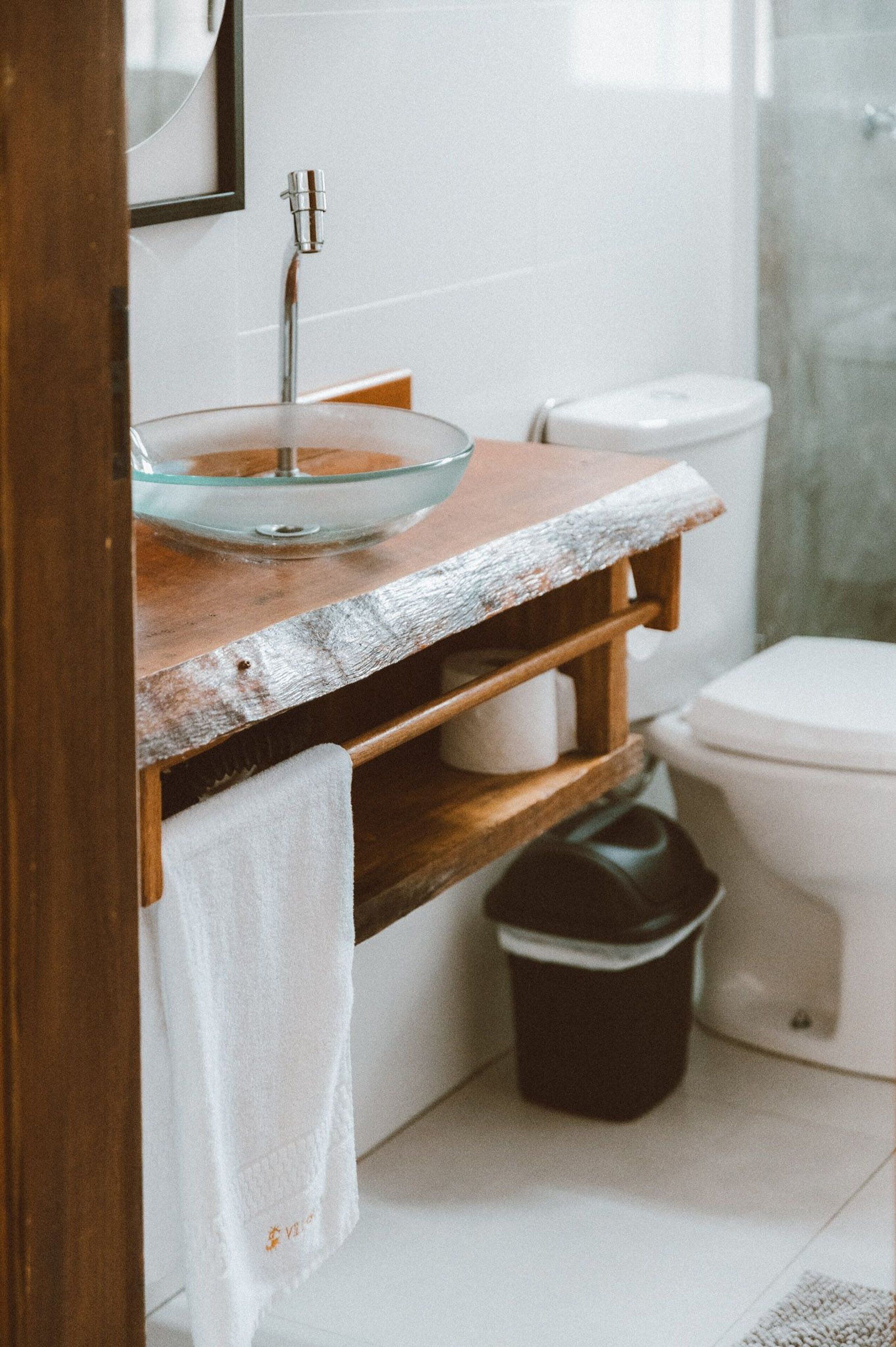 A bathroom with a glass bowl sink, wooden shelf, white toilet, and a trash bin.