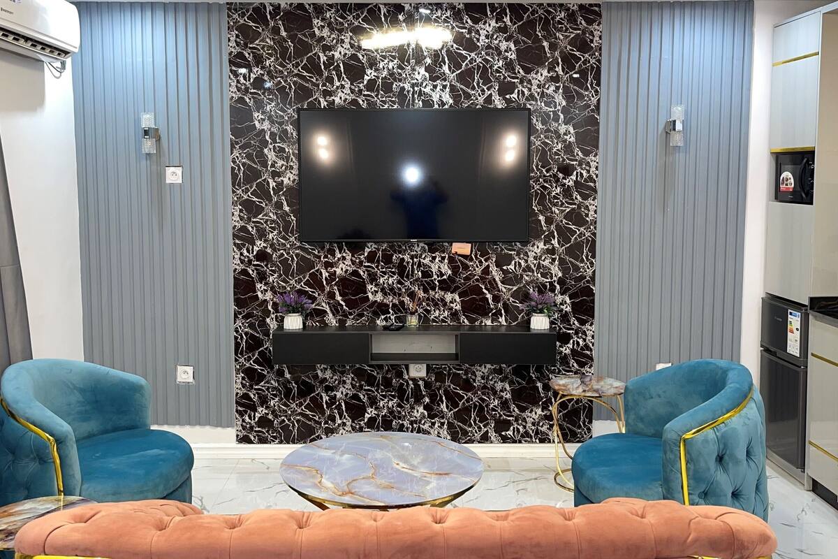 A modern living room with a large flat-screen TV mounted on a marble wall, two blue armchairs, a round coffee table, and a small side table.