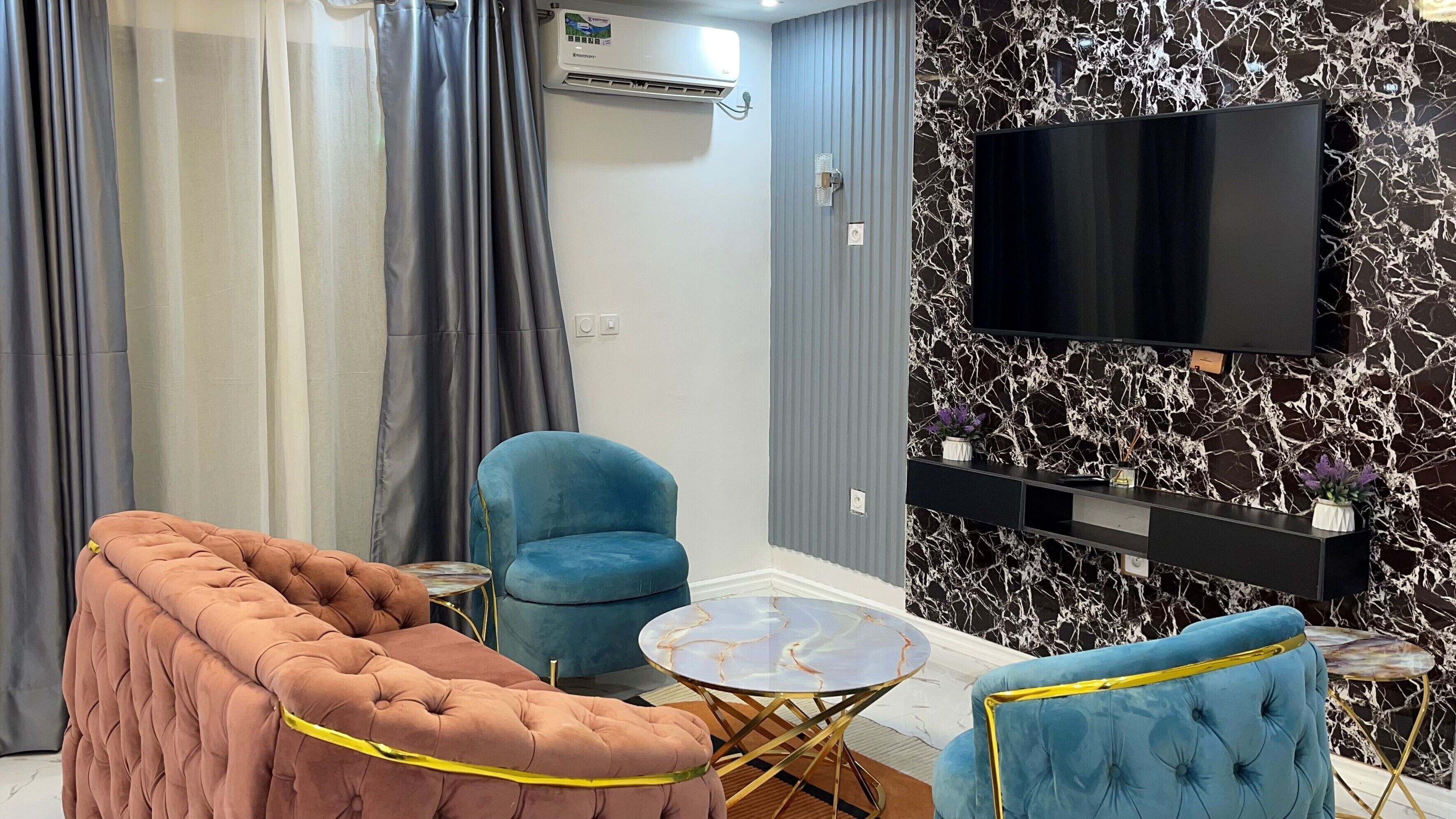 A modern living room with a tufted sofa, a blue armchair, a small round table, and a flat-screen TV mounted on a marble-patterned wall.