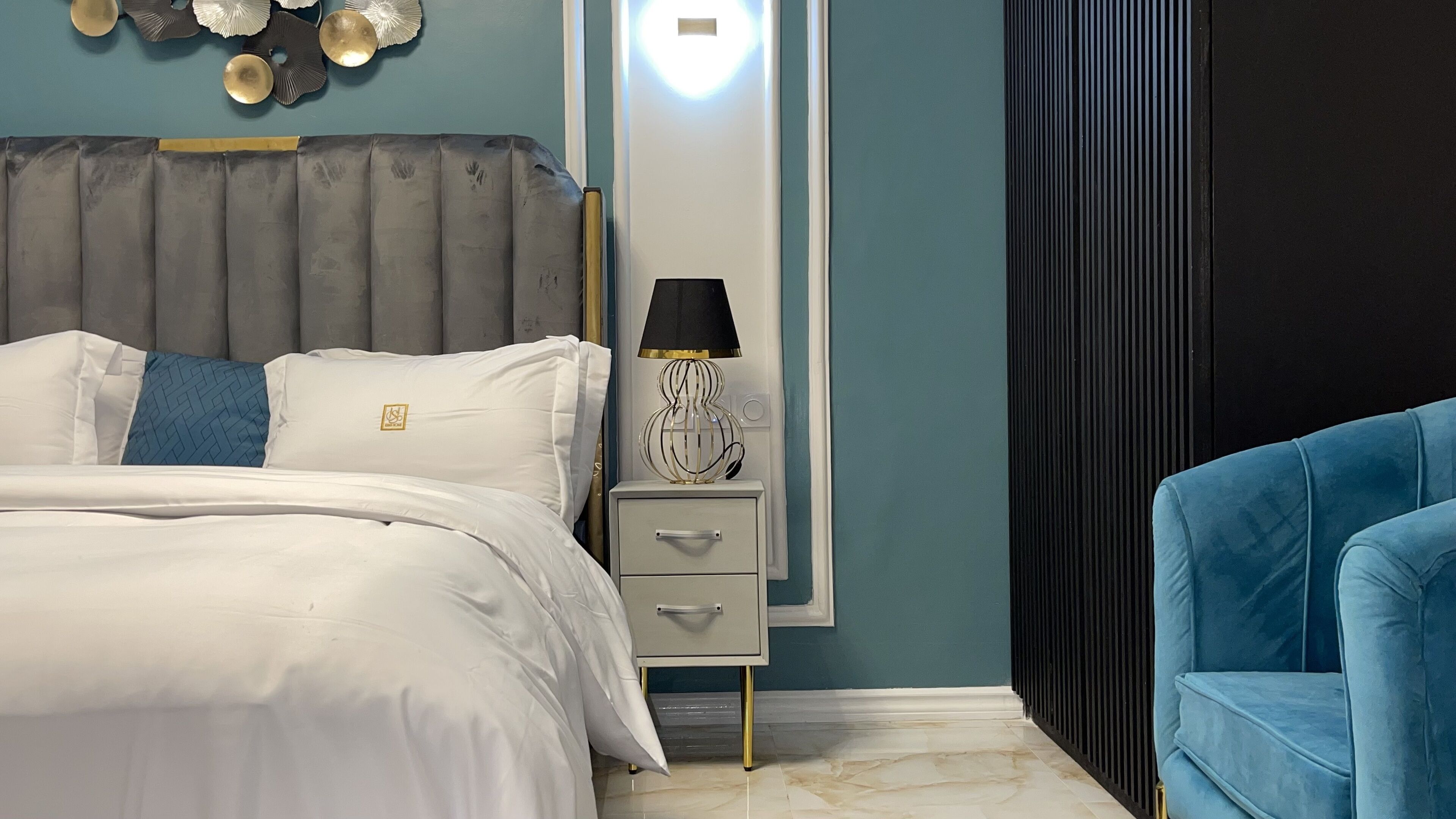Un dormitorio con cama, mesita de noche, lámpara y un sillón azul.