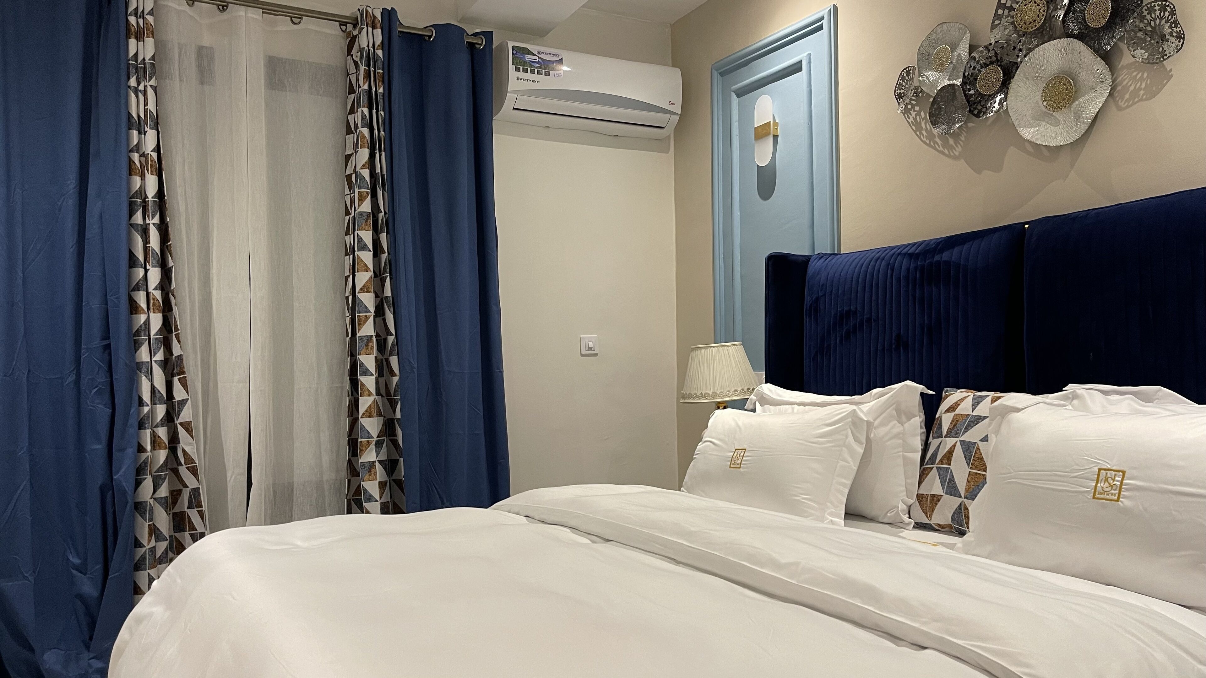 Une chambre à coucher avec un lit, des rideaux, un appareil de climatisation, une lampe et des assiettes décoratives accrochées au mur.