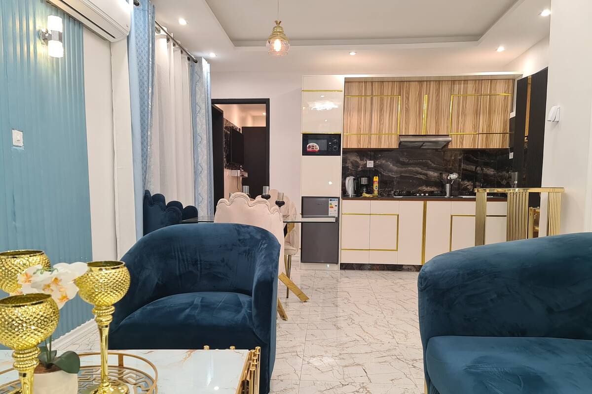 A modern living room with dark blue sofas, a marble coffee table, and a kitchen area with wooden cabinets and a black stove.