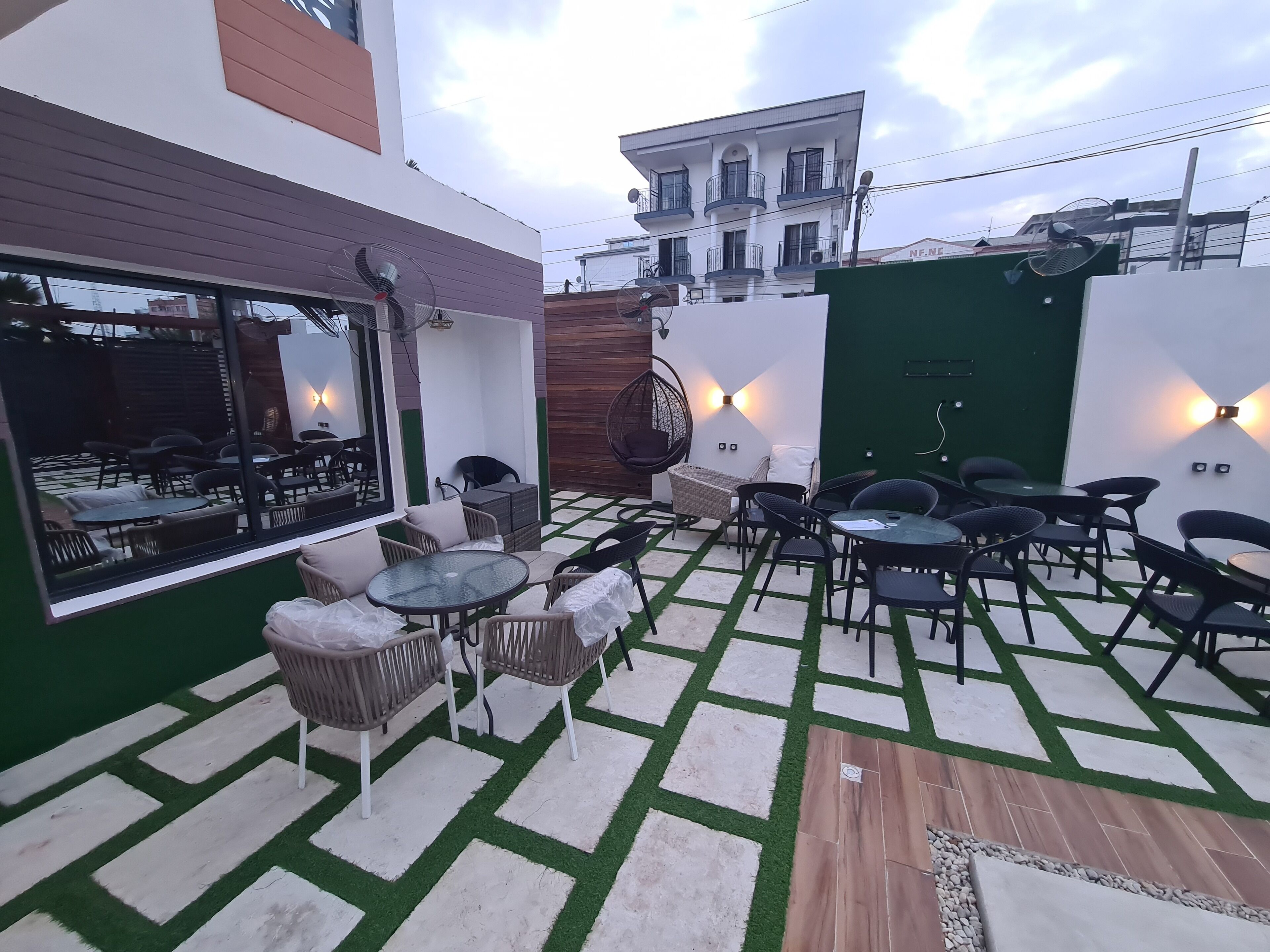 An outdoor patio area with tables and chairs, a green wall, and a view of a building in the background.