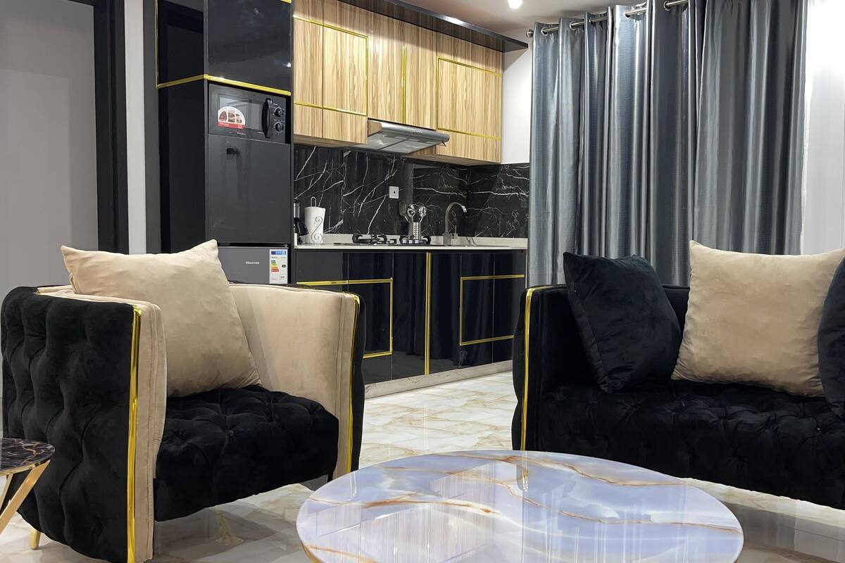 A modern living room with two black sofas with gold accents, a marble coffee table, and a kitchen in the background.