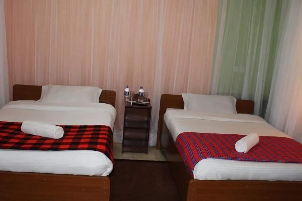 A hotel room with two single beds, each with a red and black checkered bedspread and a white pillow. There is a small table with two water bottles and glasses between the beds.