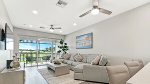 A modern living room with a grey sofa, a coffee table, and a dining table. There is a balcony with a view of the outdoors. - GreenLinks at Lely Resort · King Bed · Lake View · Golf · Sleeps 6 (Naples)