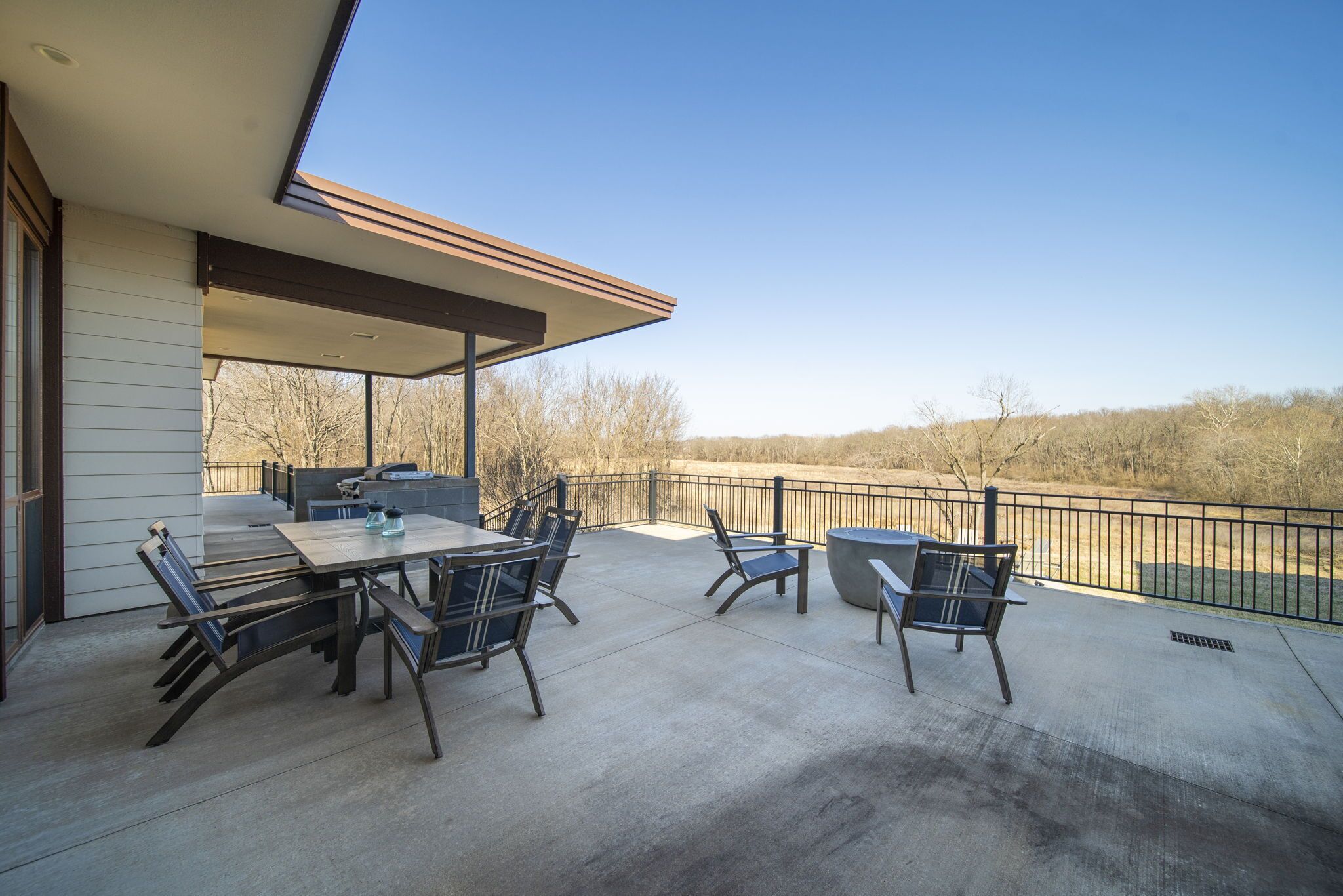 An outdoor patio with a table and chairs, a grill, and a view of a wooded area.