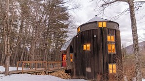 A wooden cabin with a red door and multiple windows, surrounded by trees and snow.