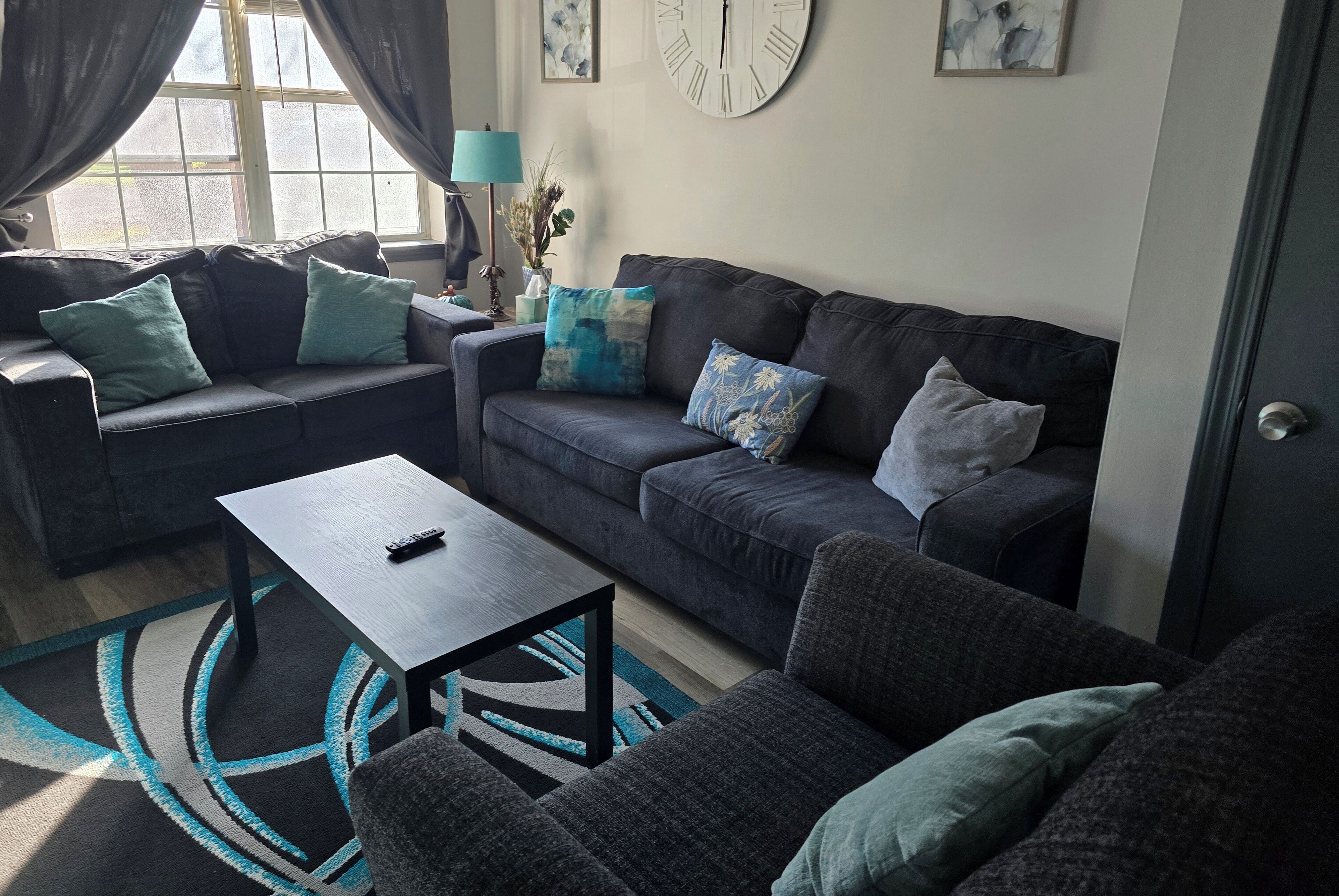 A living room with a dark grey sofa, a matching armchair, and a coffee table. There is a window with curtains, a clock on the wall, and a lamp on a side table.