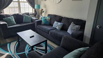 A living room with a dark grey sofa, a matching armchair, and a coffee table. There is a window with curtains, a clock on the wall, and a lamp on a side table.