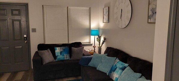 Un salon avec un canapé marron foncé, deux coussins bleus et gris, une table d’appoint avec une lampe, une horloge murale et une porte dotée d’un système de verrouillage à clavier.