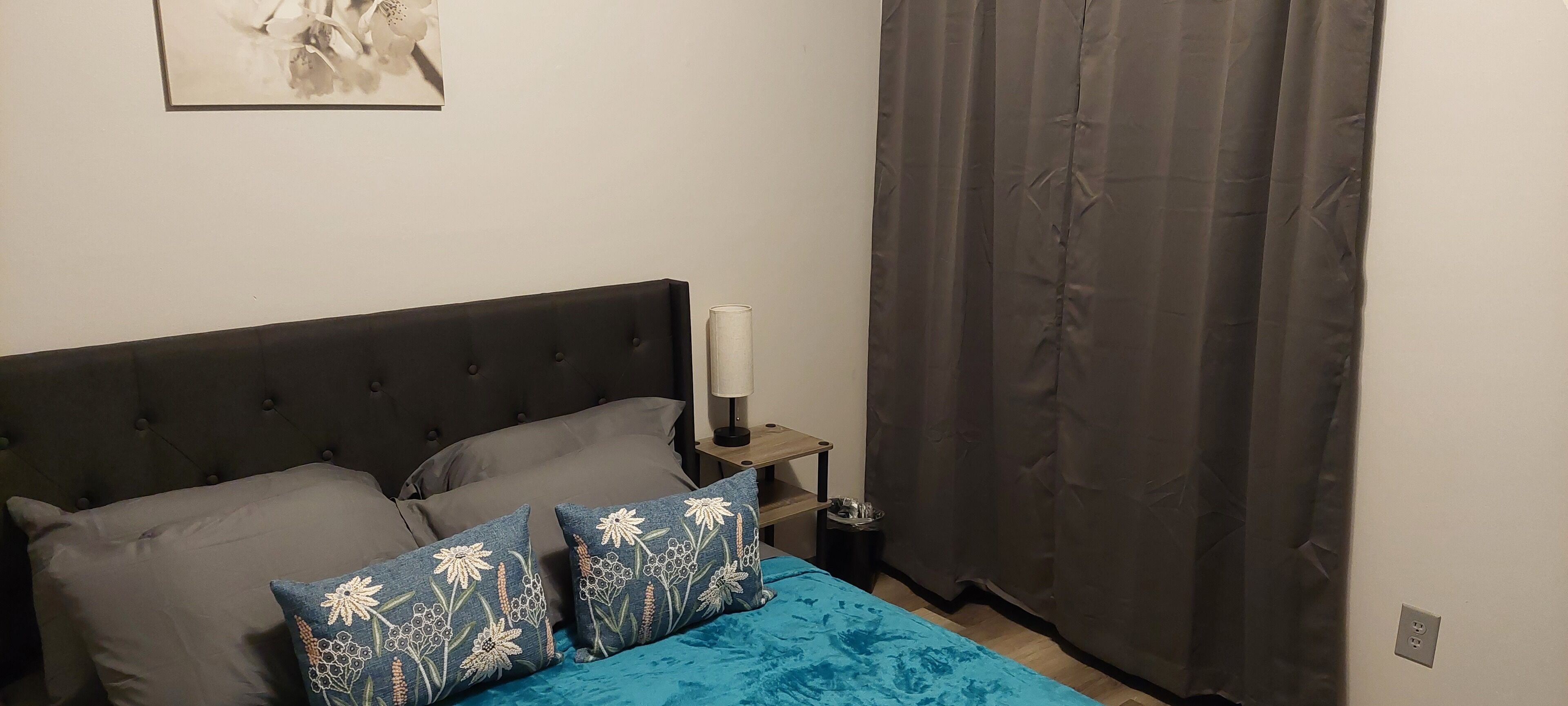 Quarto com cabeceira escura, duas almofadas cinzentas, colcha azul e cortina cinza.