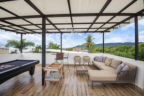 A covered outdoor area with a pool table, seating, and a view of greenery and hills.