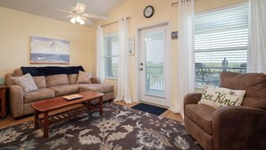 A living room with a beige sofa, a brown armchair, a wooden coffee table, and a floral rug. There is a ceiling fan, a painting of a boat on the water, and a clock on the wall. - Nature's Landing C302 Pool and Amazing Water View! (Cedar Key)
