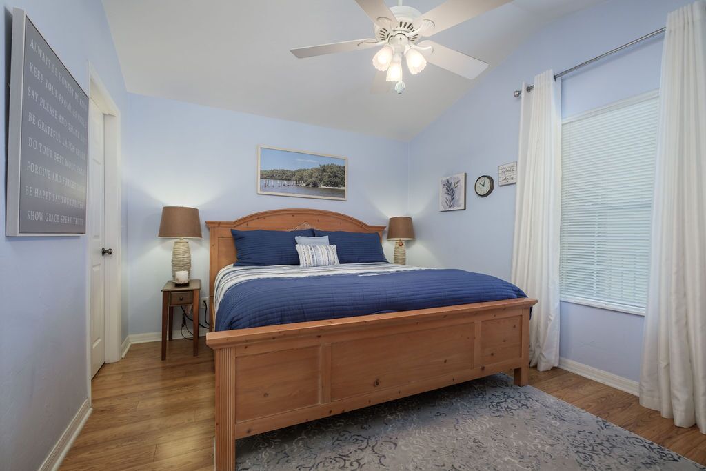 Une chambre avec un lit en bois, deux lampes de chevet, un ventilateur de plafond, un tableau encadré au mur et une fenêtre avec des rideaux.