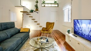 A modern living room with a sofa, a round glass coffee table, a golden armchair, a TV, and a staircase with a plant shelf.