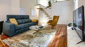 A modern living room with a blue sofa, a glass coffee table, a patterned rug, a wooden floor, a TV, and a staircase with a plant shelf.