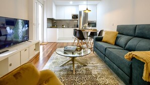 A modern living room with a sofa, a coffee table, and a TV. There is a dining area with a table and chairs. The kitchen is visible in the background with white cabinets and a stainless steel range hood.