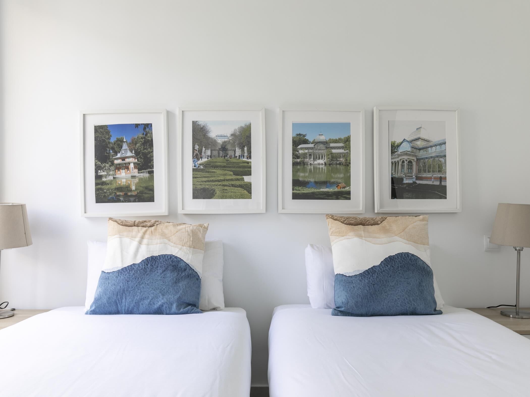 A hotel room with two beds, each with a white bedspread and blue pillows. There are four framed pictures on the wall depicting various landscapes and buildings.