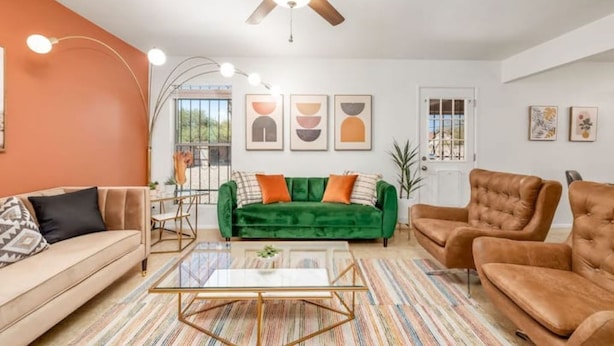 Un salon avec un canapé vert, un canapé beige et un fauteuil marron. On y trouve un ventilateur de plafond, une table basse en verre et un tapis à rayures.