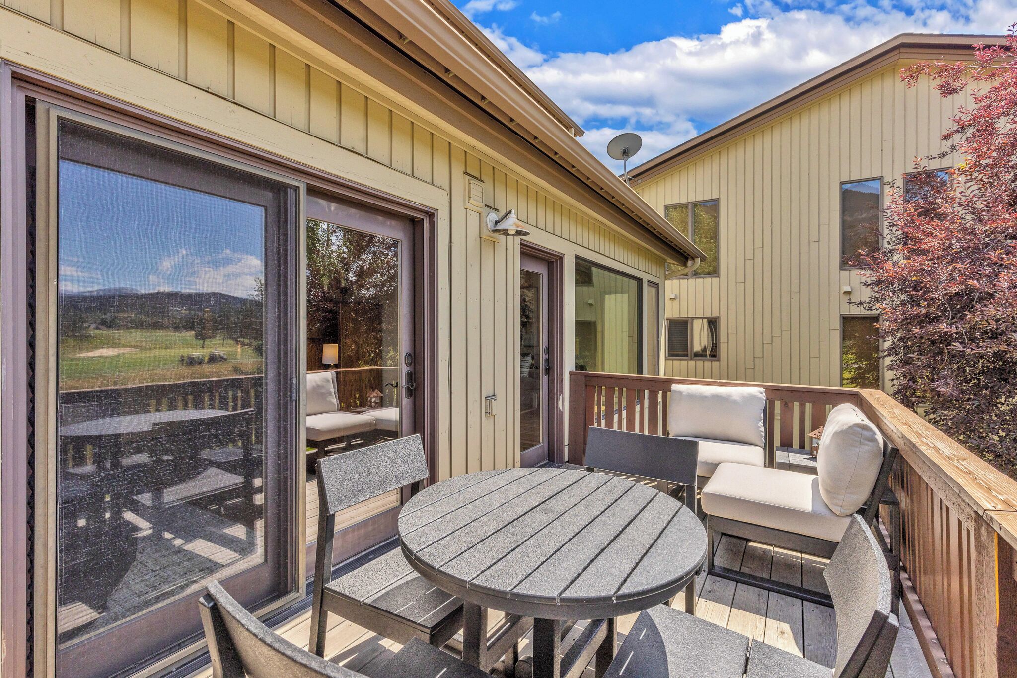 A covered outdoor patio with a round table and chairs, a sliding glass door leading to a deck, and a view of a green landscape.