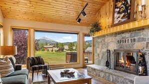 A cozy living room with a stone fireplace, large windows offering a view of the outdoors, and a comfortable seating area. - Lone Peak Lookout l Big Sky Golf Course (Big Sky)